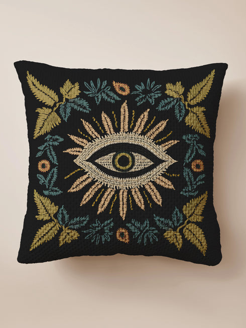 A black woven throw pillow featuring intricate embroidery of an Evil Eye motif in gold, ivory, and green, surrounded by detailed botanical flourishes.